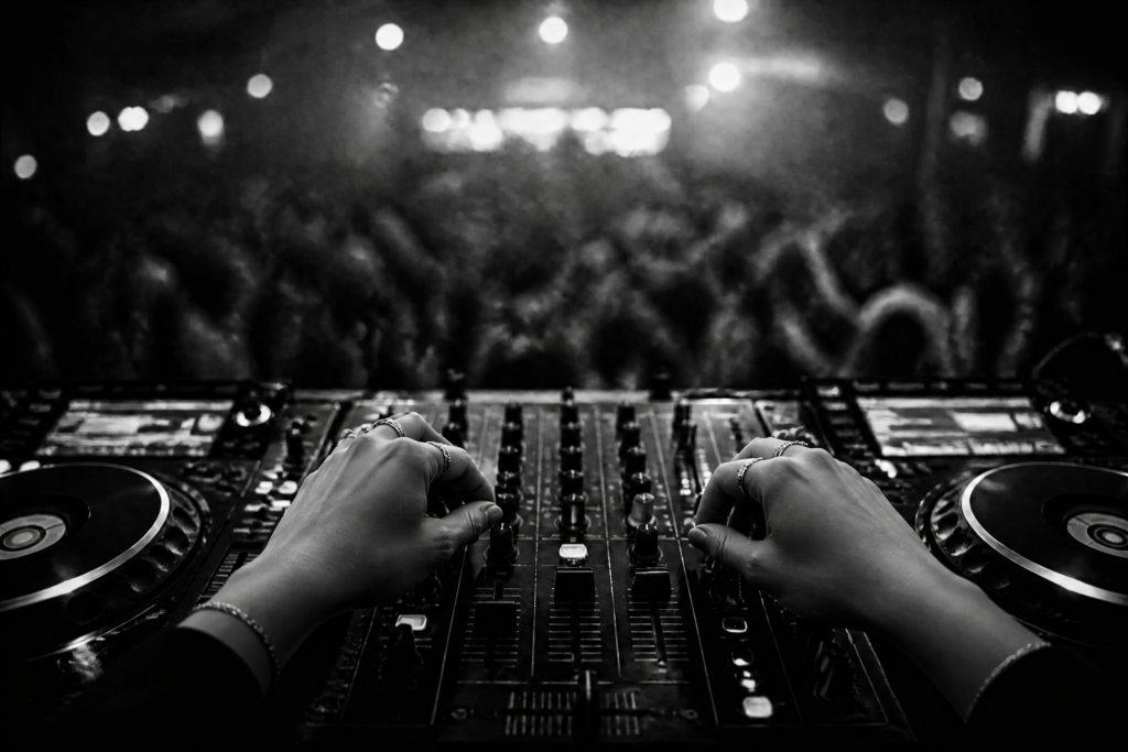 Nahaufnahme eines DJ-Pults von hinten, Hände an den Reglern, Crowd unscharf im Hintergrund.